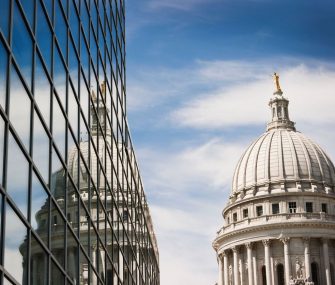 Horizontal view of the Wisconsin State Capitol Dome reflecting into the windows of a steel and glass office building on a sunny day.