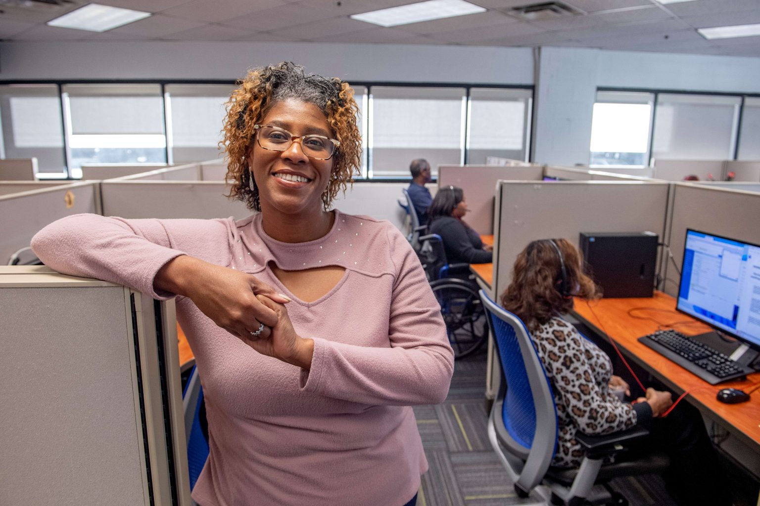 Lighthouse Enterprises contact center agent smiling at her workstation with colleagues working behind her