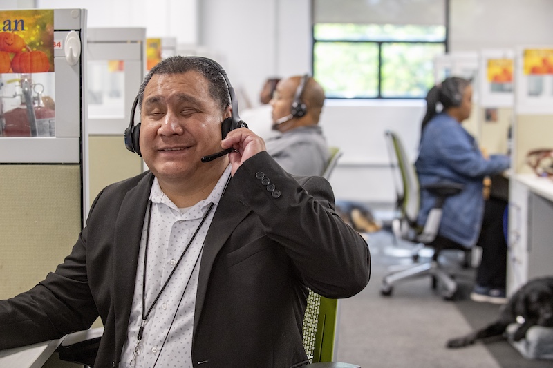 Call center agent wearing a headset and smiling at his desk in an open-plan office with coworkers in the background