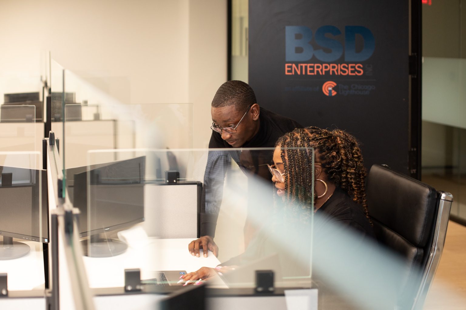 Two coworkers at a desk with computers behind glass partitions in a modern office; a BSD Enterprises sign is on the wall behind them.
