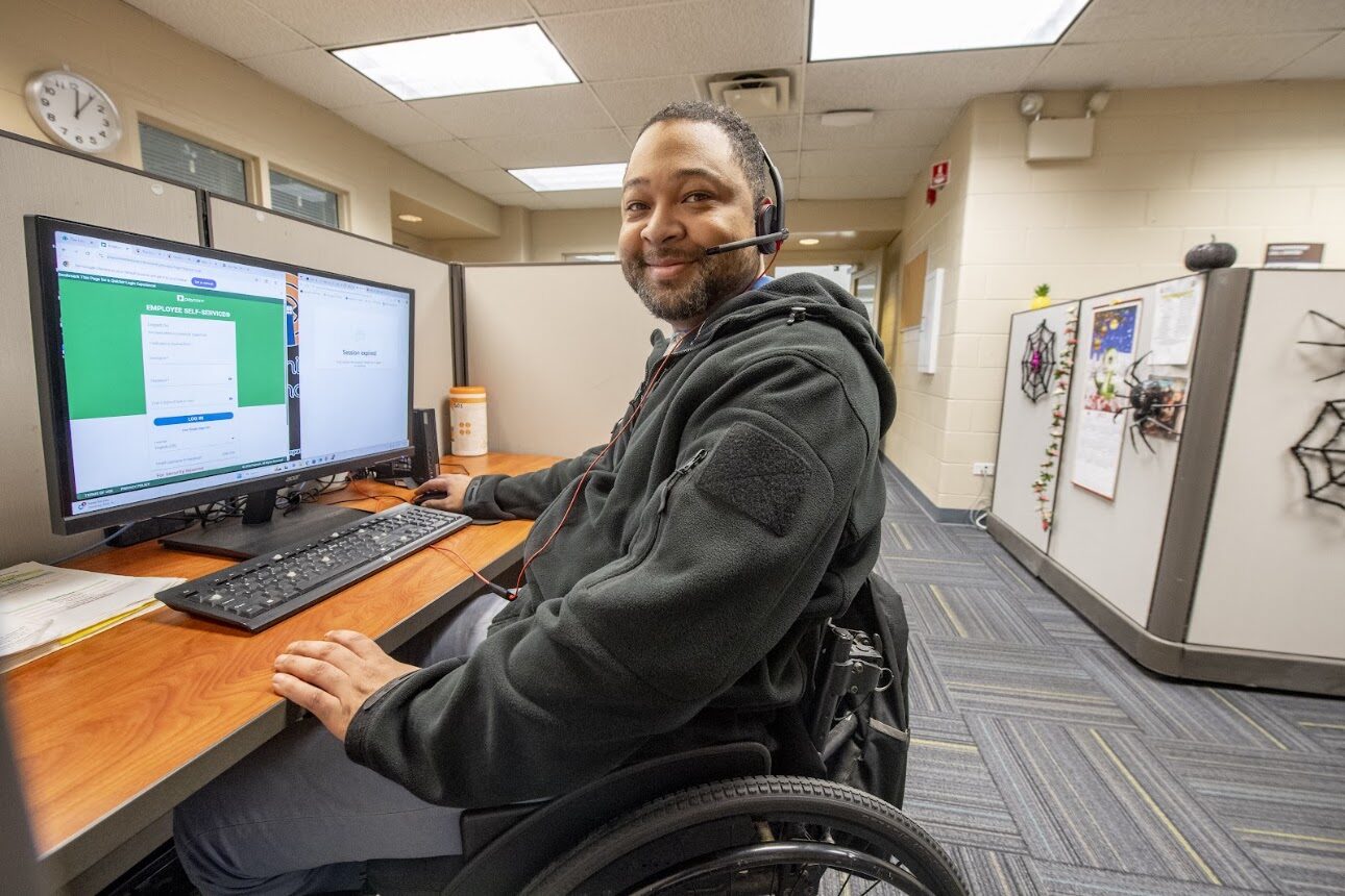 A Lighthouse Enterprises contact center agent who uses a wheelchair works at a dual-monitor workstation while wearing a headset.