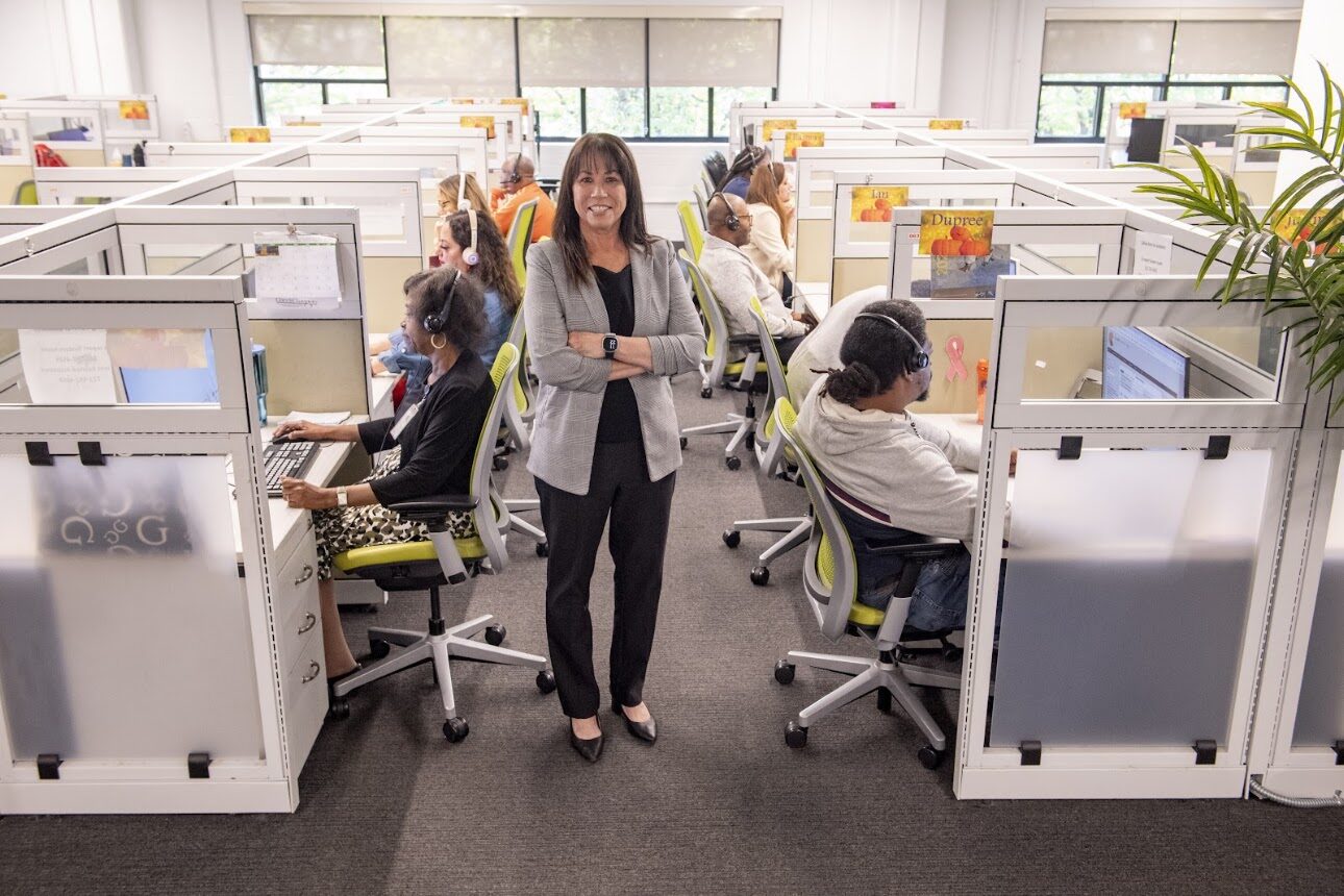 A bustling Lighthouse call center with rows of cubicles; a smiling woman in a gray blazer stands in the center among seated agents wearing headsets.